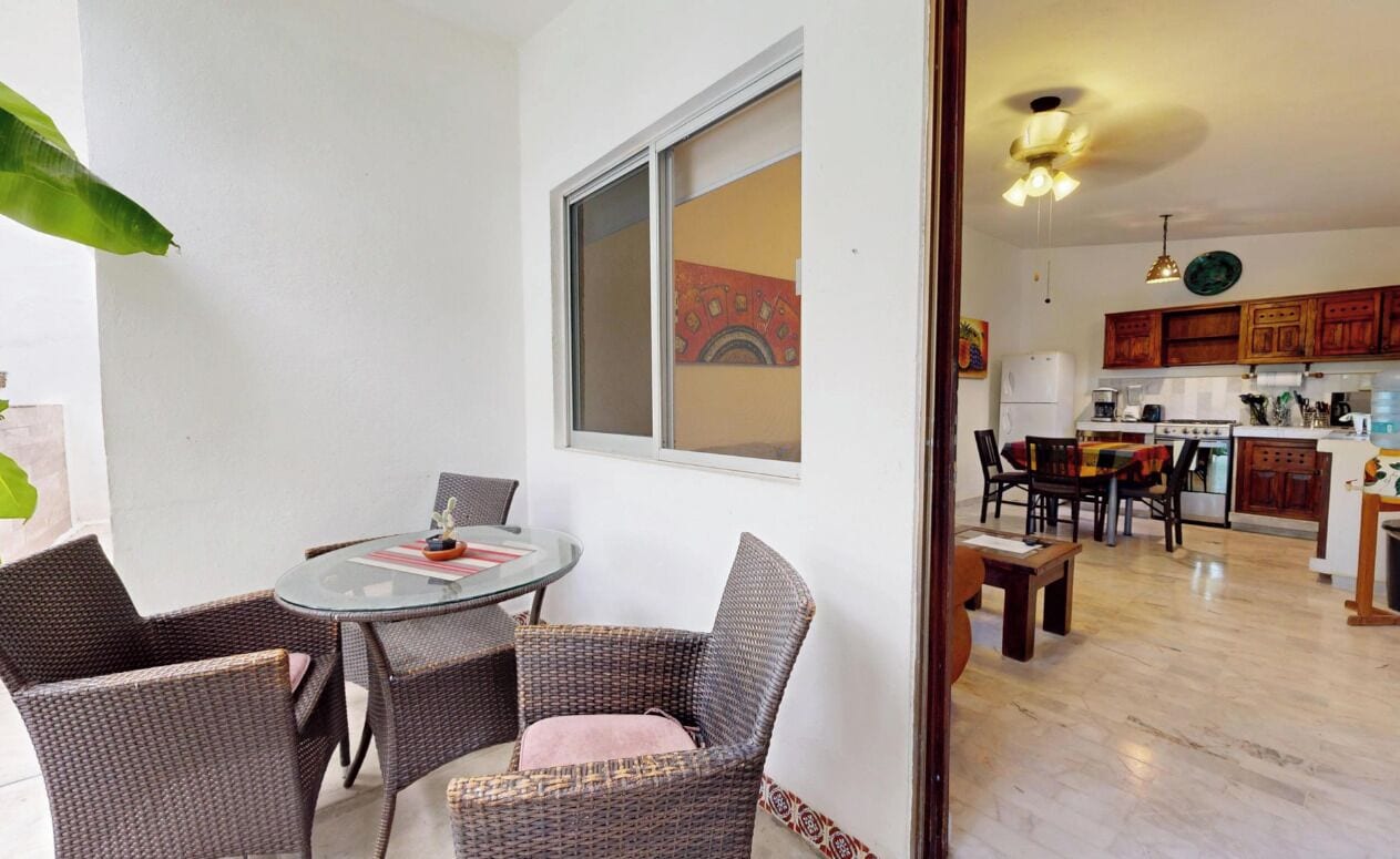 A dining area with wicker chairs and a glass table, a kitchen with wooden cabinets, and a view through a window with a decorative painting.