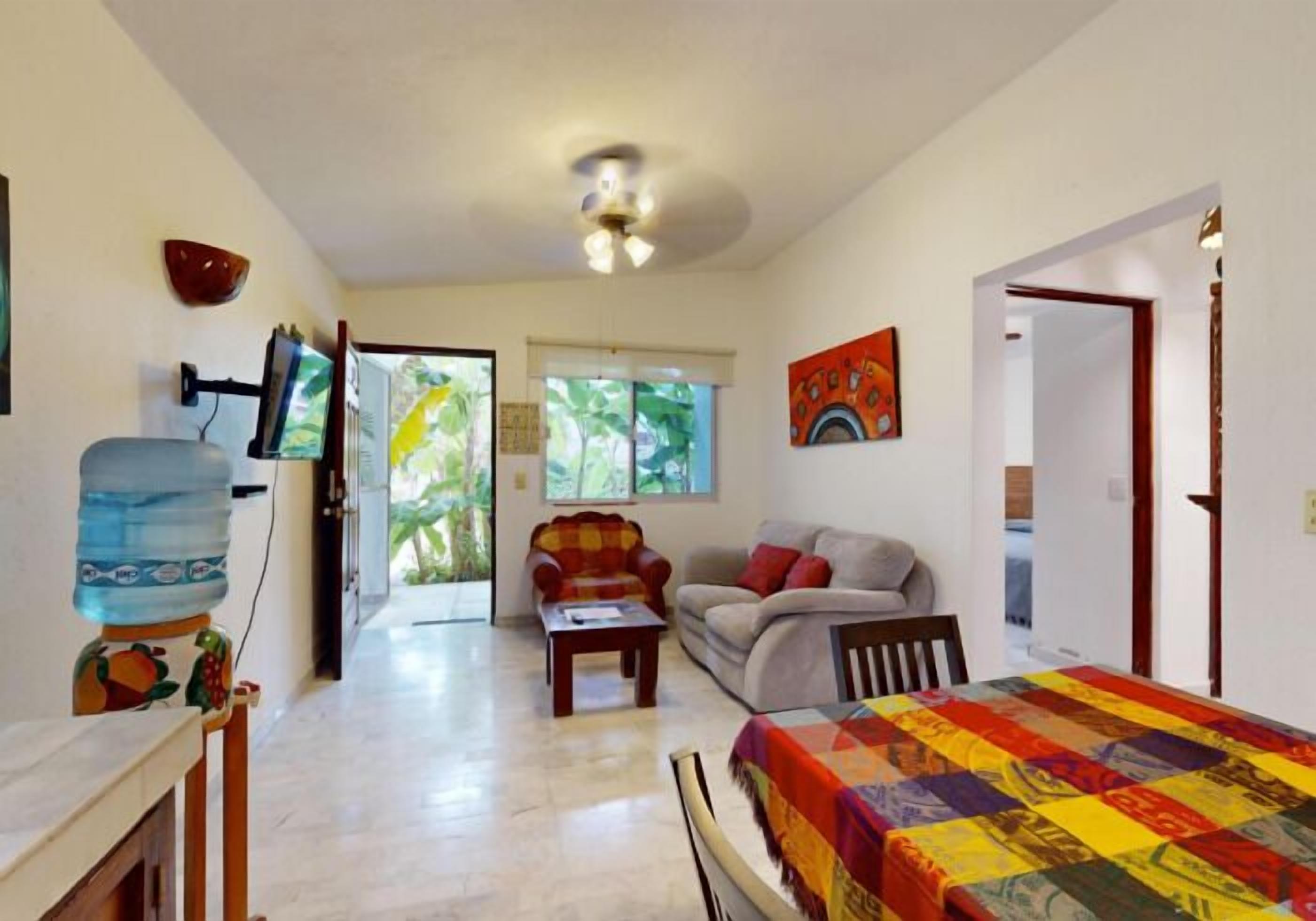 A room with a bed, a sofa, a chair, a table, and a water dispenser. There is a window with a view of greenery outside.