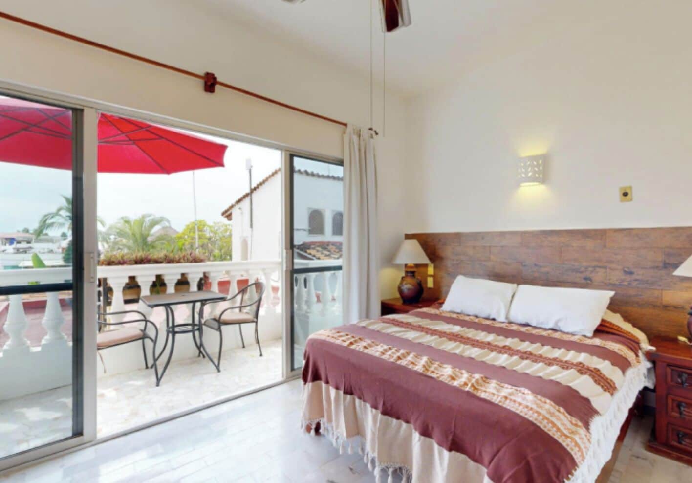 Habitación de hotel con balcón, cama con colcha estampada, mesita de noche con lámpara y vistas al exterior.