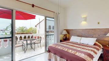 Habitación de hotel con balcón, cama con colcha estampada, mesita de noche con lámpara y vistas al exterior.