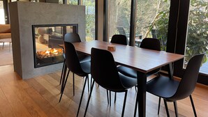 Un comedor moderno con mesa de madera y sillas negras, chimenea y ventanas amplias con vista a árboles.