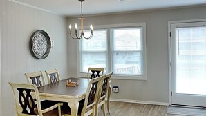 A dining room with a wooden table and chairs, a chandelier, and two windows with blinds. - You-N-Sea by Topsail Realty (Holly Ridge)