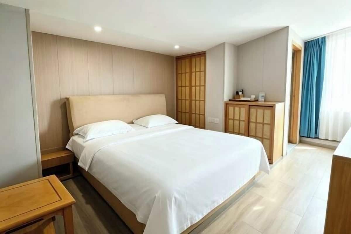 Una habitación de hotel con una cama grande, cabecera de madera, una mesita de noche de madera, un armario de madera y vistas al exterior a través de una ventana con cortinas.