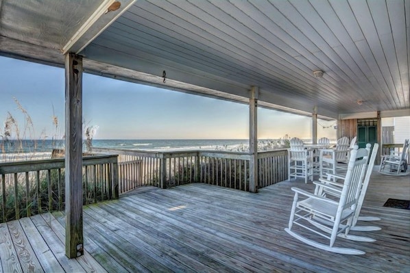 A wooden deck with white rocking chairs and tables, overlooking the ocean.