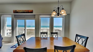 Un comedor con una gran mesa de madera y seis sillas, ventanas amplias con vista a la playa y una araña.