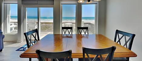 A dining room with a large wooden table and six chairs, large windows with a view of the beach, and a chandelier.