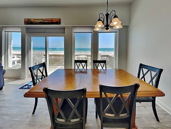 Una sala da pranzo con un grande tavolo in legno e sei sedie, ampie finestre vista spiaggia e un lampadario.