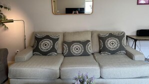 Un salon avec un canapé gris, trois coussins à motifs, un miroir fixé au mur et une petite table sur laquelle se trouve un haut-parleur.