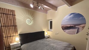 A bedroom with a bed, a nightstand, a wall-mounted lamp, a window, and a beach mural.