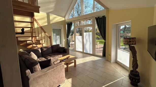 A spacious living room with a brown leather sofa, a wooden coffee table, and a TV on the right. A staircase leads to an upper level, and there is a large sliding glass door to the outside.