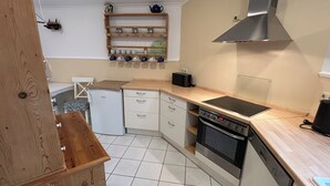 A compact kitchen with a built-in oven, a stovetop, and a microwave. There is a wooden shelf above the sink holding cups and a kettle.