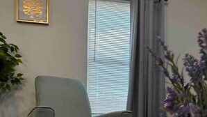 A room with a grey armchair, a white table, a window with blinds, and a framed picture on the wall. - Lemon Home - 2 Queen BR (Edinburg)