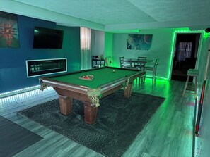 Een pooltafel met groen vilt, een biljarthouder en keuen. Er is een televisie aan de muur bevestigd en een klein tafeltje met stoelen.