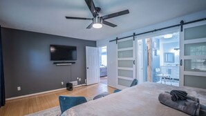 Une chambre moderne équipée d’un lit, d’un ventilateur de plafond, d’un téléviseur à écran plat et d’une porte-fenêtre coulissante donnant sur un balcon.