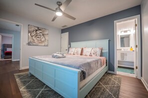 Une chambre avec un lit bleu, un ventilateur de plafond, une grande œuvre d’art florale et une salle de bain visible à travers une porte ouverte.