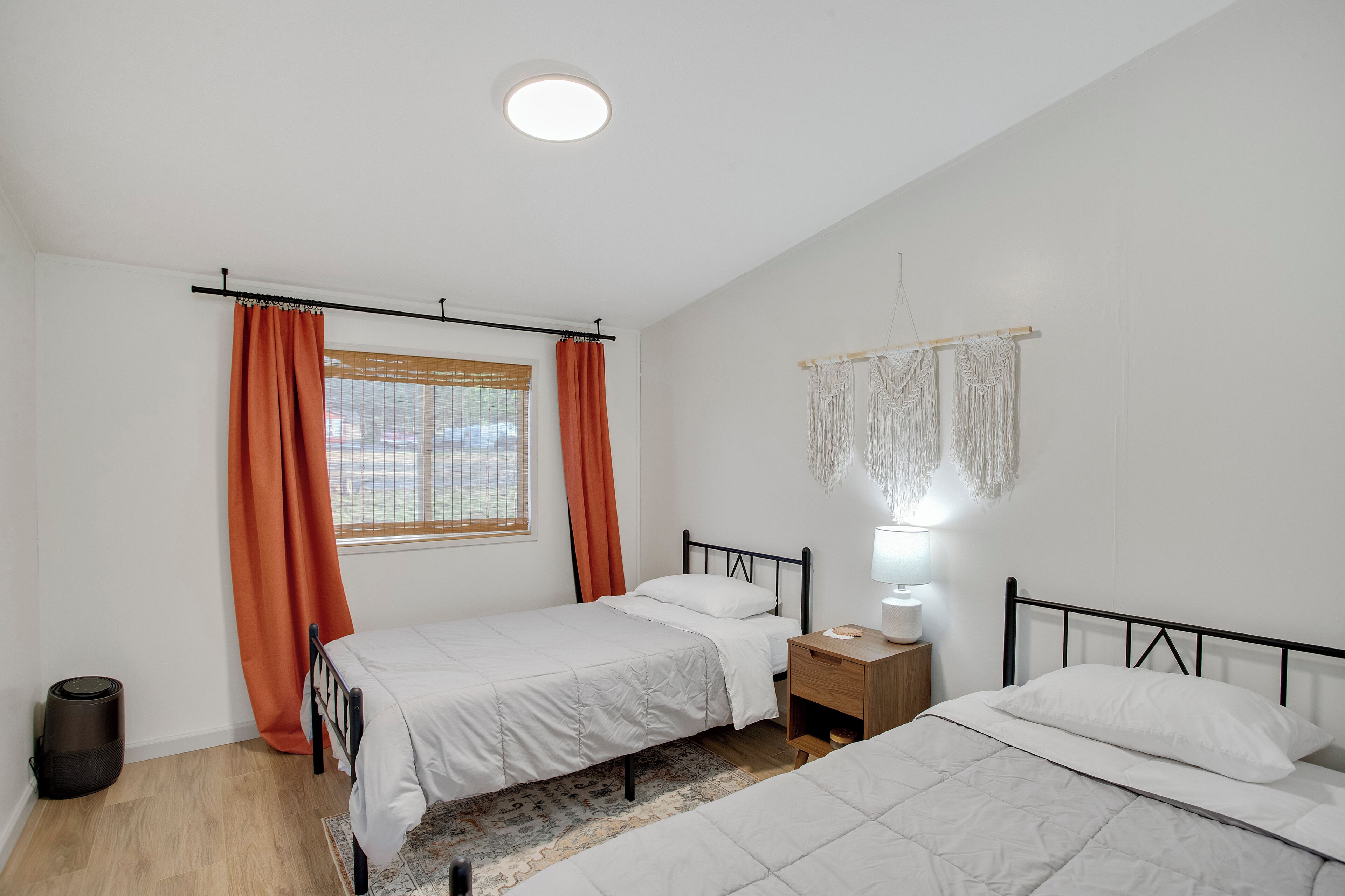 Ein Schlafzimmer mit zwei Betten, einem Fenster mit orangefarbenen Vorhängen, einem Nachttisch mit Lampe und einem Wandbehang aus Makramee.