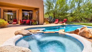 A swimming pool with a hot tub, surrounded by rocks and red chairs, with a building and patio area in the background. - Troon North Private Pool & Spa (Scottsdale)
