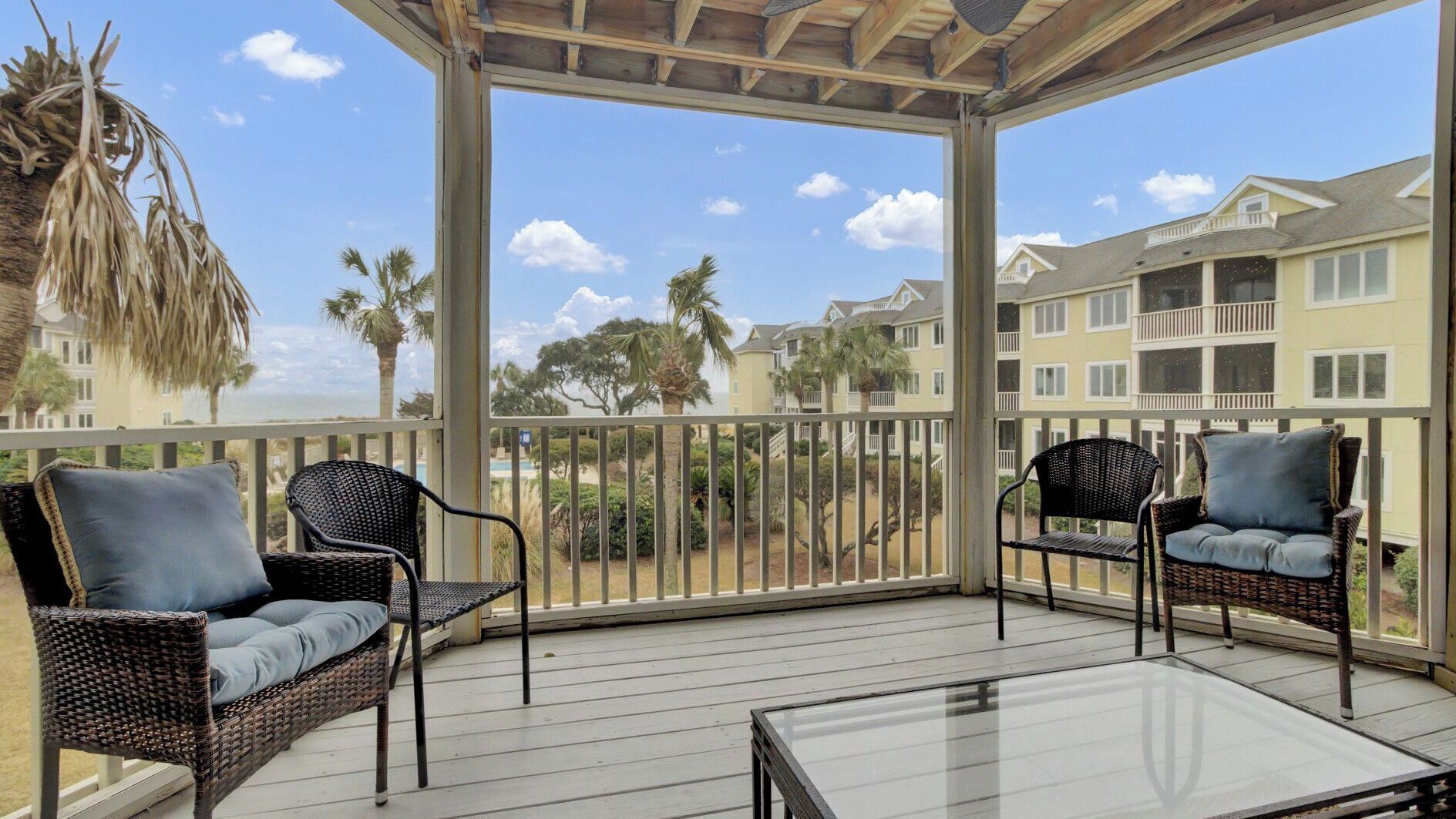 A covered outdoor area with wicker furniture and a glass table, overlooking a residential area with houses and palm trees.