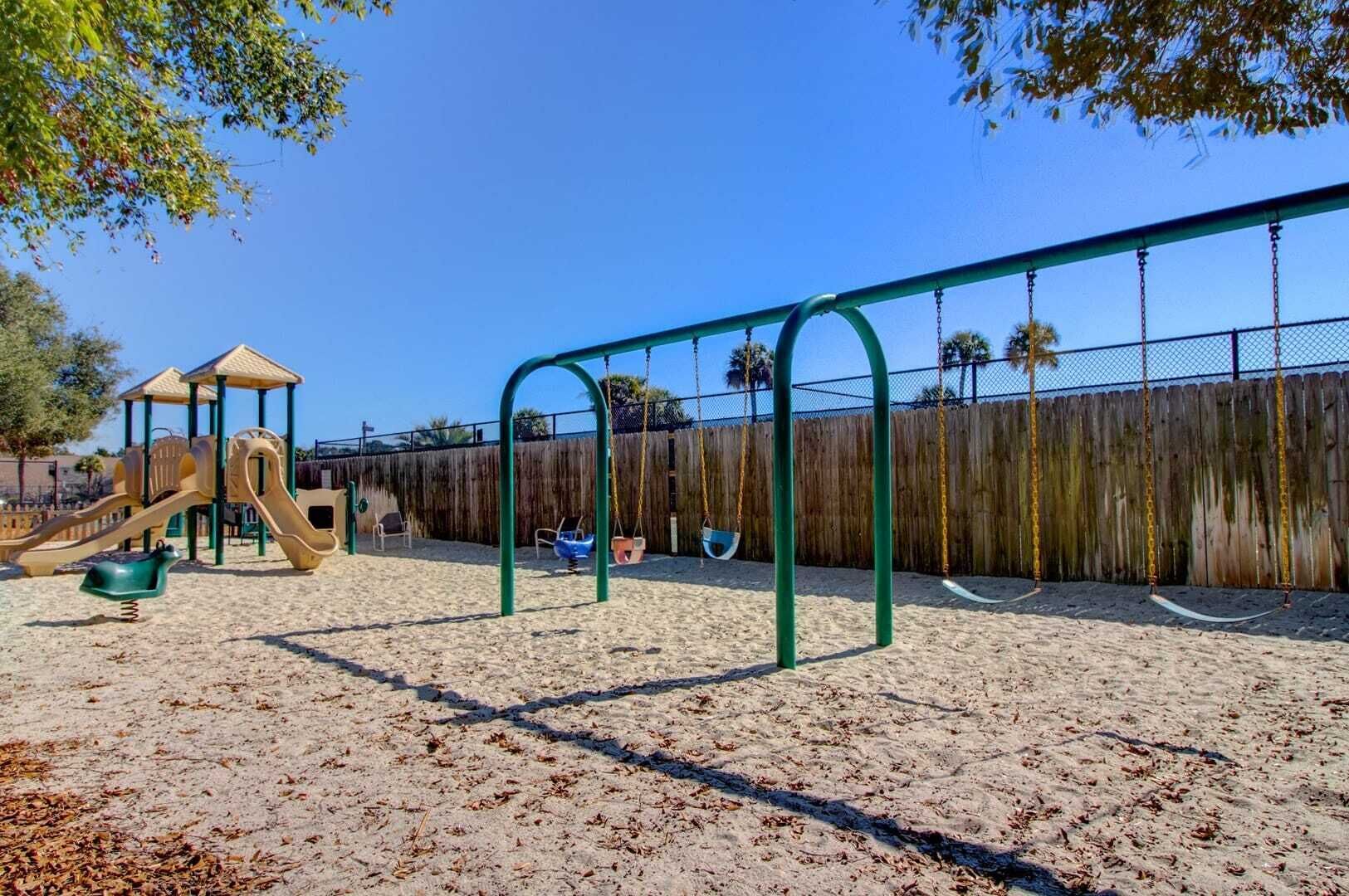 A playground with swings and a slide, surrounded by a wooden fence and trees.
