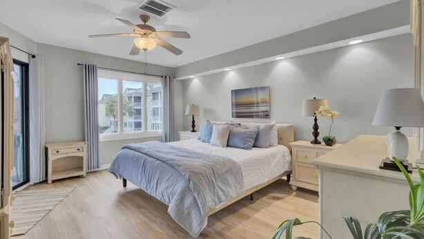 A bedroom with a bed, bedside tables, a ceiling fan, a window, and a view of buildings outside. - 202 D Port O Call by Avantstay Beach Condo (Isle of Palms)