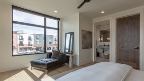 A modern bedroom with a large window, a bed, a sofa, and a bathroom with a wooden door.