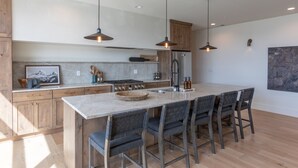 A modern kitchen with a large island, pendant lighting, wooden cabinets, and a stainless steel refrigerator.