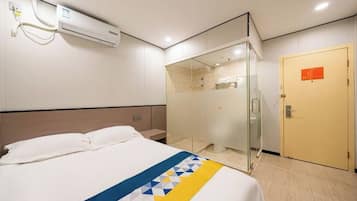 A hotel room with a bed, a glass shower enclosure, an air conditioning unit, and a door with a sign.