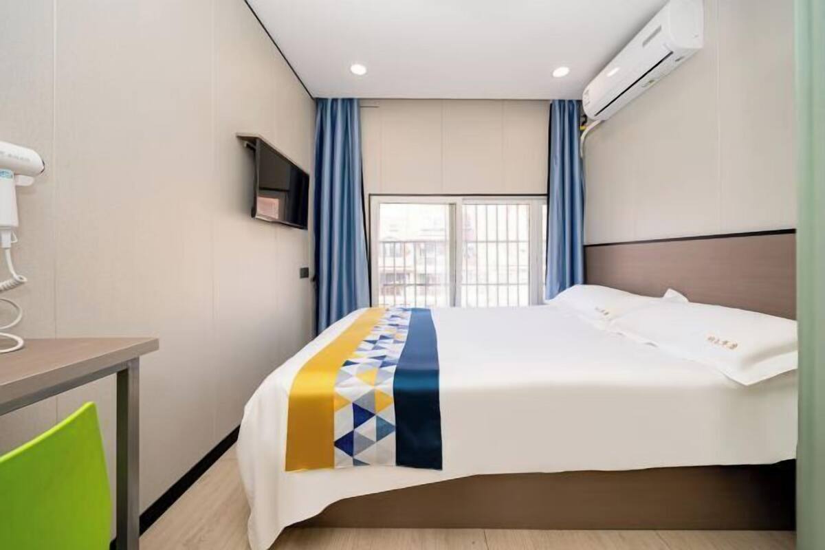 A hotel room with a bed, a desk, a chair, an air conditioning unit, and a window with curtains.