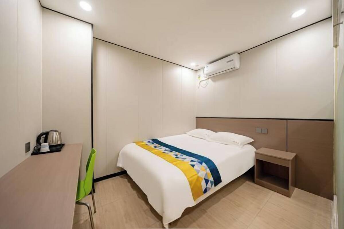 A hotel room with a bed, bedside table, chair, and a small table with a kettle.