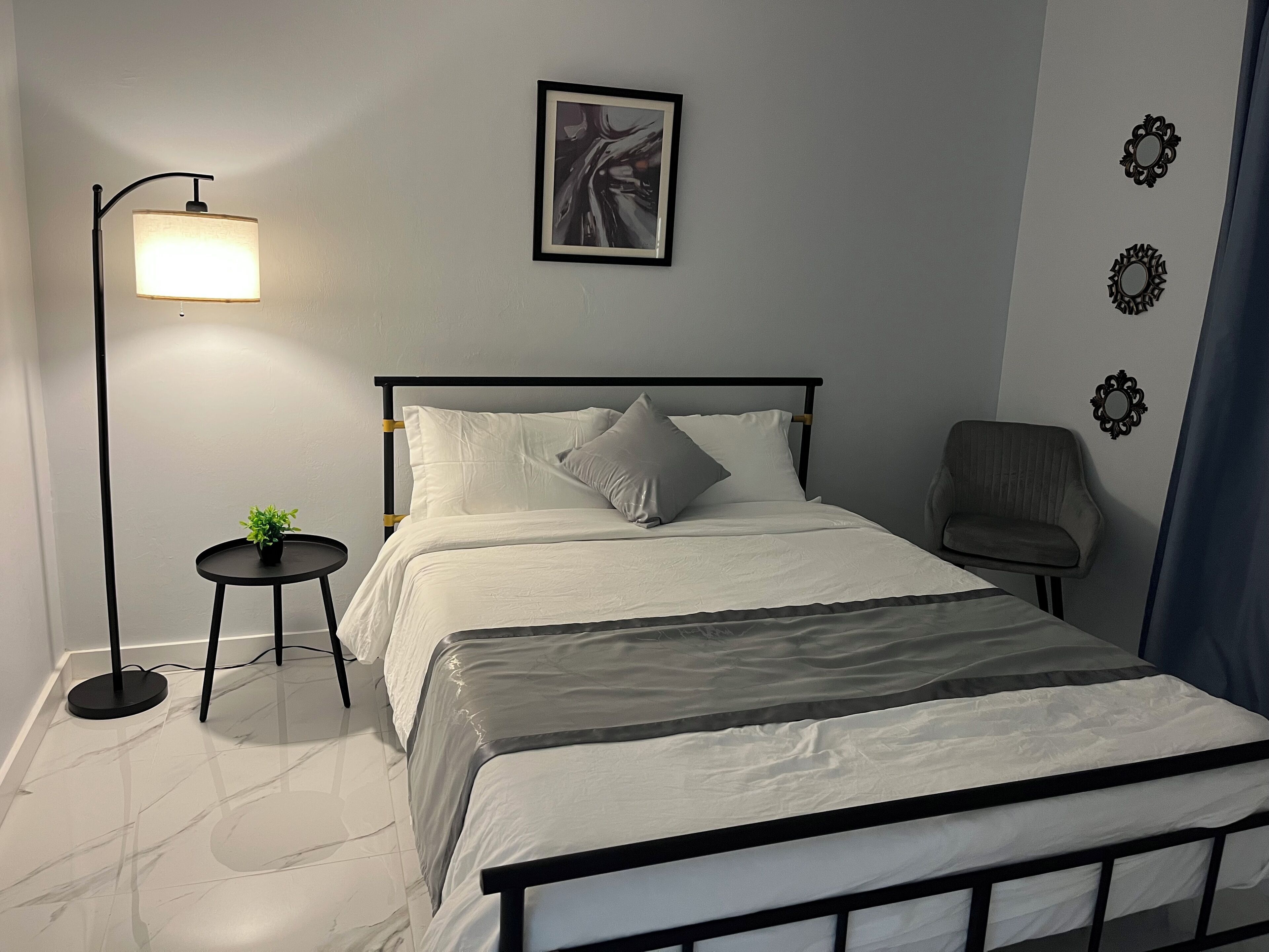 Une chambre à coucher bien rangée, avec un lit, une table de chevet, une lampe, une chaise et un tableau encadré accroché au mur.