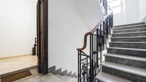 Eine Treppe mit hölzernem Geländer und schwarzen Metallstreben, die zu einer Zwischenplattform mit hellem Boden und einem kleinen dekorativen Gegenstand links führt.