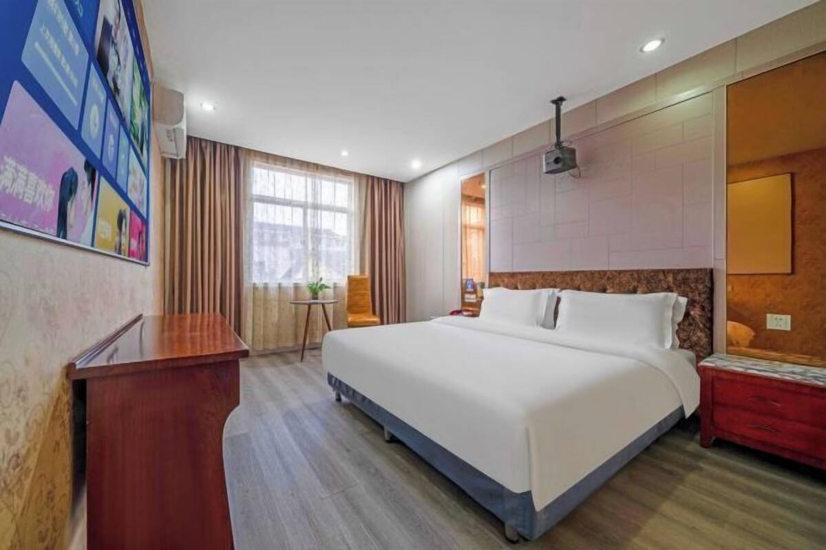 A hotel room with a large bed, a desk, a chair, a window with curtains, and a wall-mounted projector.