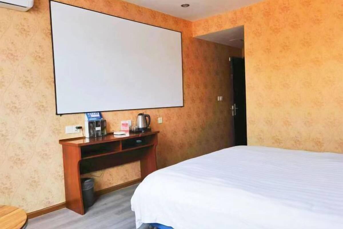 A hotel room with a bed, a desk with a coffee maker, and a large blank screen on the wall.