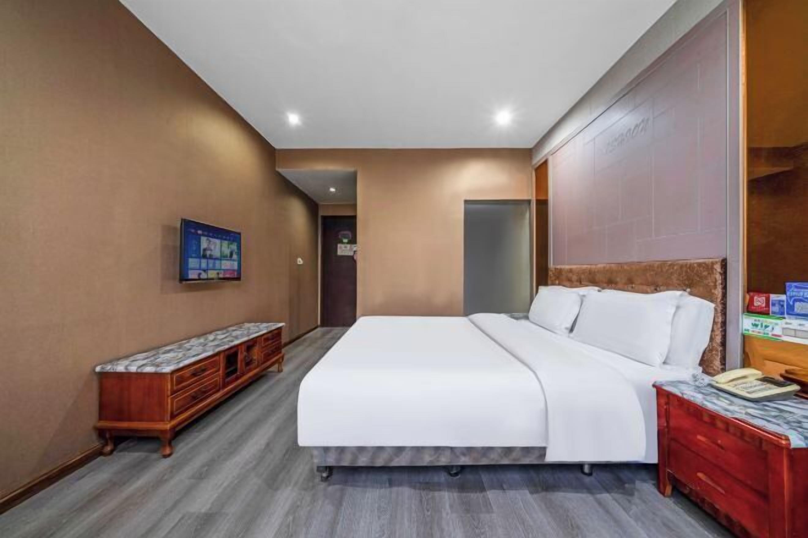 A hotel room with a bed, a wooden bench, a nightstand, and a wall-mounted TV.