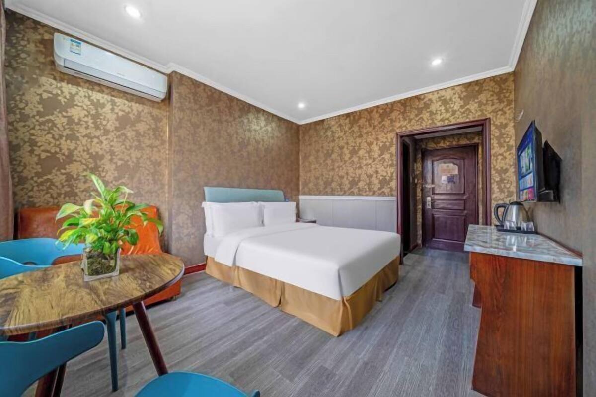 A hotel room with a bed, a small table with chairs, a potted plant, a TV, and a wooden desk.