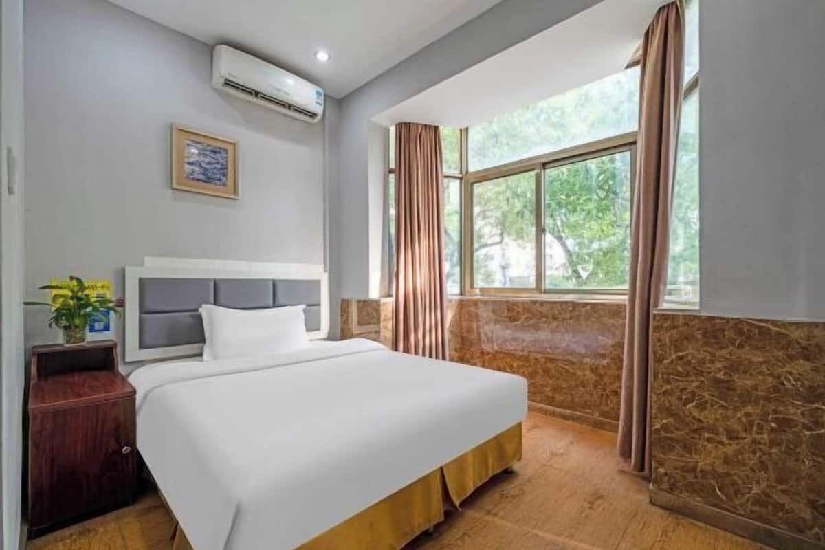 A hotel room with a bed, an air conditioner, a nightstand, a window with curtains, and a view of greenery outside.