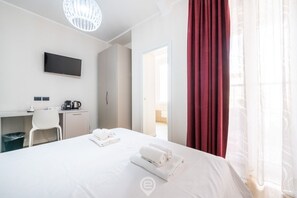 A hotel room with a bed, a desk with a flat-screen TV, a coffee maker, and a view of the outside. - Estay Piazza Repubblica - Suite Mirto (Cagliari)