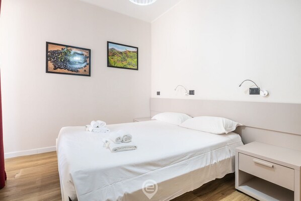 A clean, modern hotel room with a neatly made bed, two framed pictures on the wall, a bedside table with a lamp, and a small drawer unit. - Estay Piazza Repubblica - Suite Mirto (Cagliari)