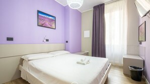 Ein Hotelzimmer mit einem Bett, einem Bild an der Wand, einem Fenster mit Vorhängen und einem Papierkorb.