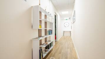 A hallway with white walls, a wooden floor, and white shelving units with books. There is a fire exit sign on the wall and a clock on the right side.