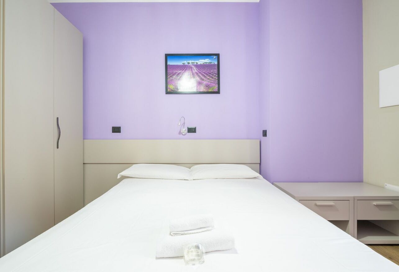 A neatly made bed with white linens and a folded towel, a bedside table, and a wall-mounted picture of a lavender field.