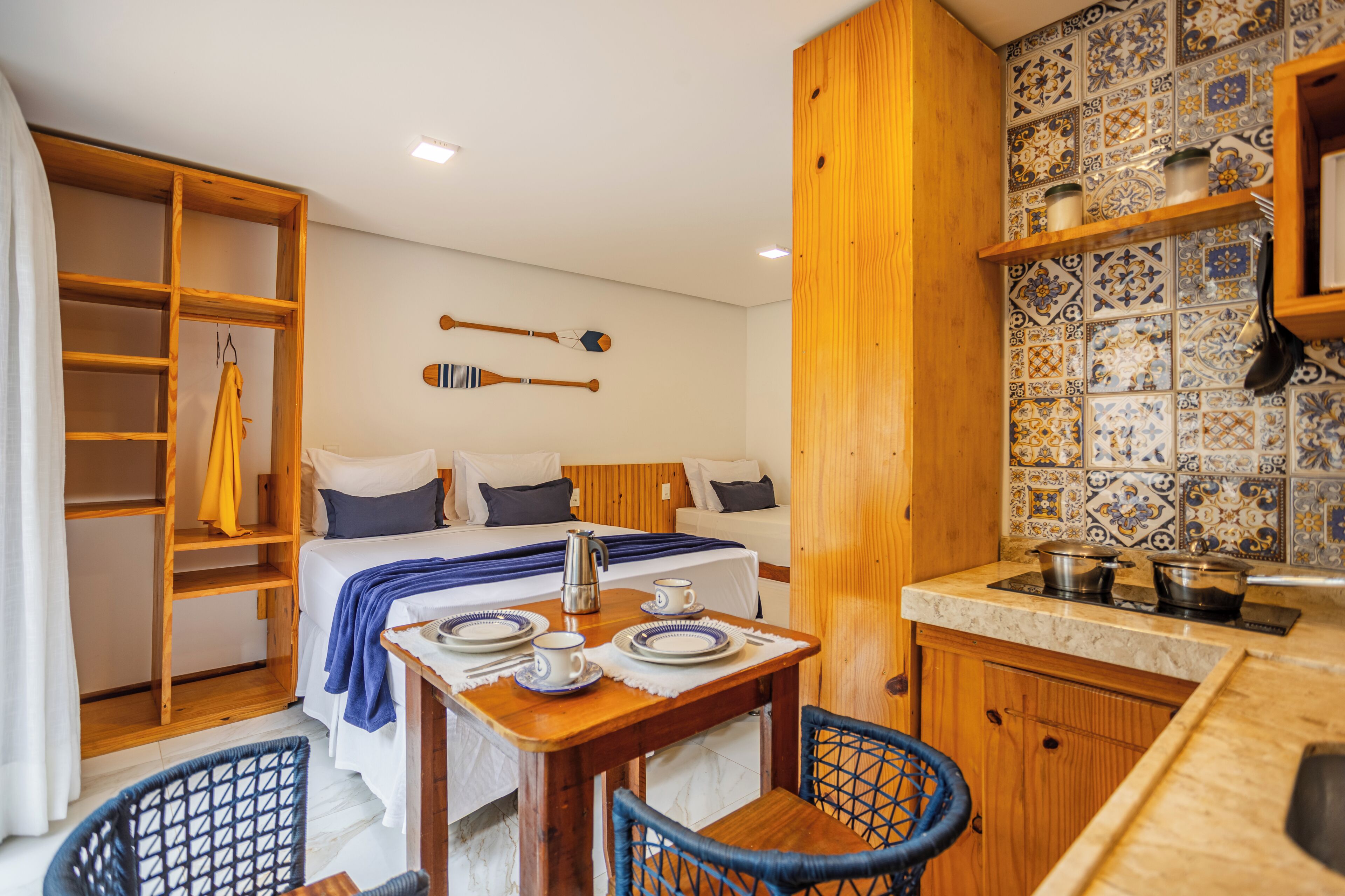 A compact hotel room with a bed, a small dining table with chairs, a kitchenette, and a wooden wardrobe.