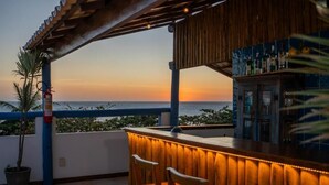 Un bar con vista al océano al atardecer, sillas de madera y barandilla azul.