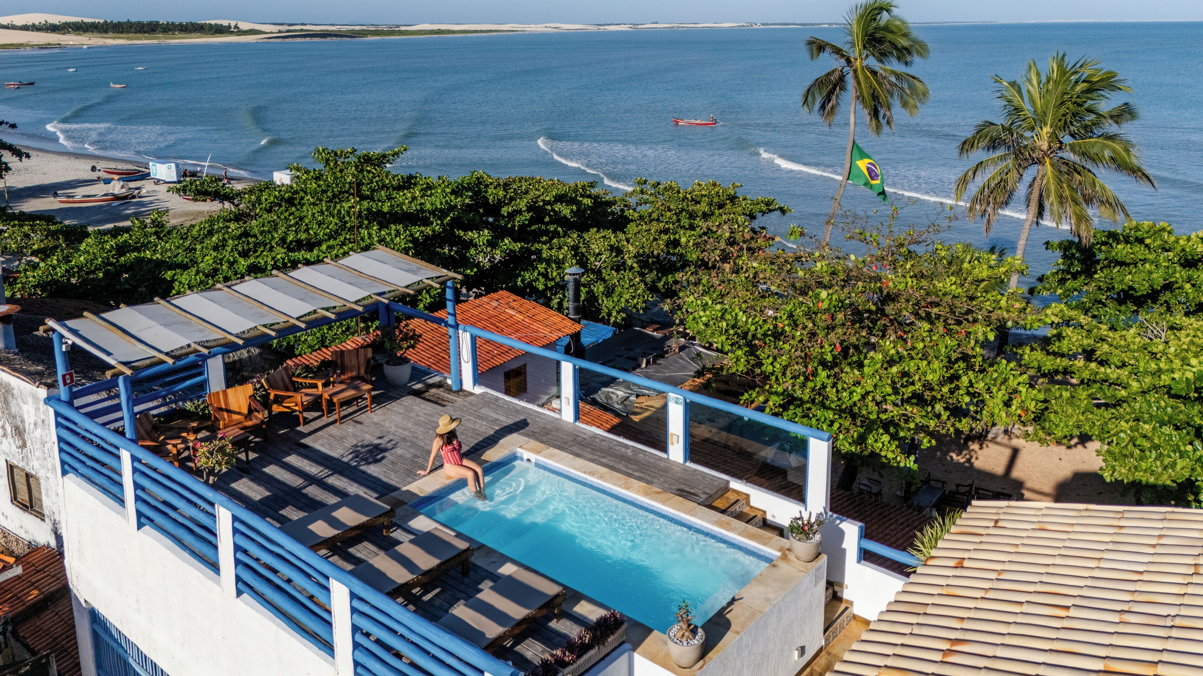 A rooftop pool with a view of the sea, surrounded by greenery and a building with a tiled roof.
