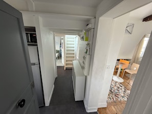 Um corredor estreito com escada que leva a um quarto. Uma cozinha é visível à esquerda, e uma sala de jantar com mesa e cadeiras à direita.