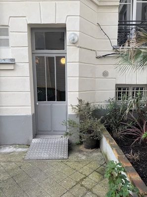 Entrada de um edifício com uma porta cinzenta, um degrau que a precede e um vaso de planta ao lado da porta.