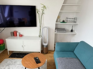 Uma sala de estar com televisão, um armário branco, um sofá azul, uma mesa de centro de madeira e um ventilador.