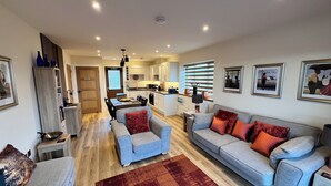 A modern living room with a grey sofa, a blue armchair, and a red rug. There is a kitchen in the background with white cabinets and a dining area.
