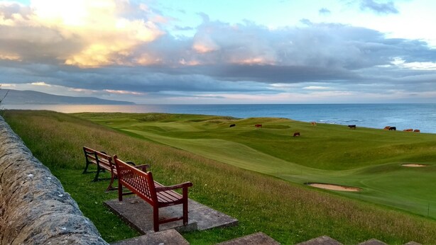 A golf course with a view of the sea, a bench, and a stone wall.