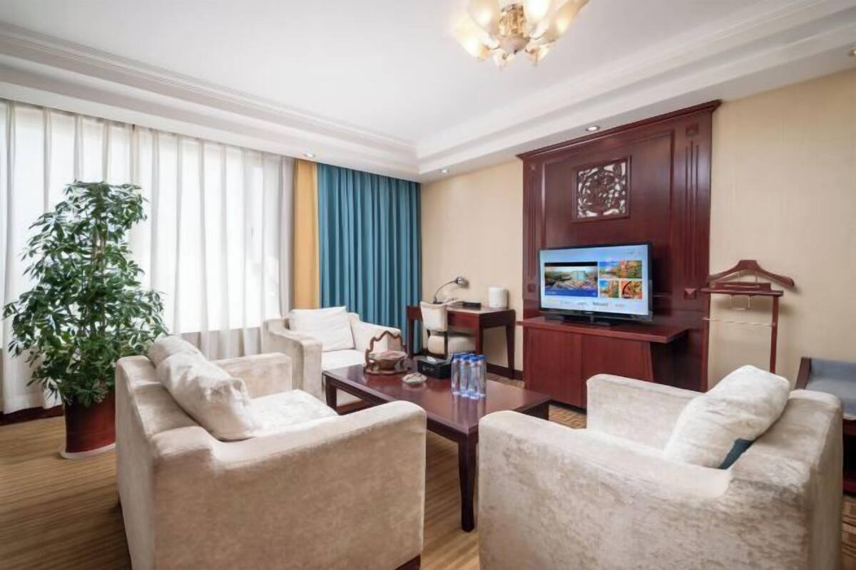 A hotel room with a sofa set, a television, a desk, and a potted plant.